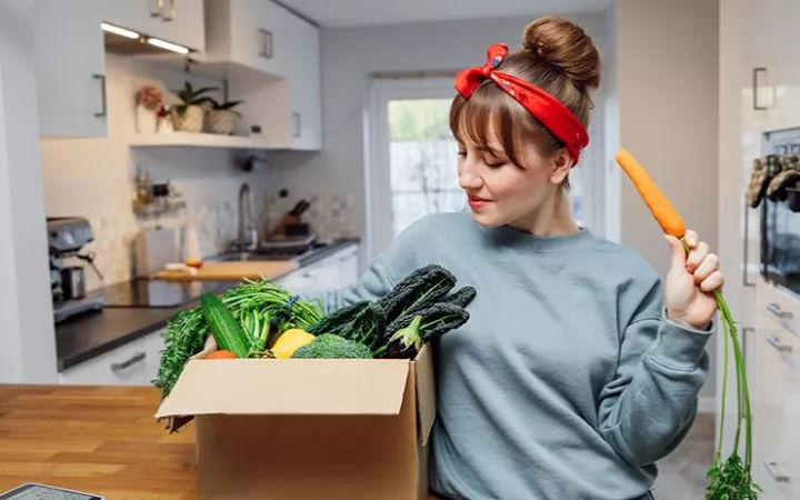 Frau mit einer Kiste voller Gemüse & einer Karotte in der Hand
