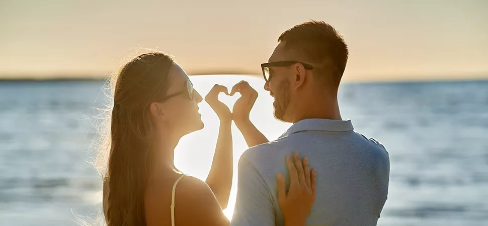 Ein Paar am Strand blicken in den Sonnenuntergang und formen ihre Hände zu einem Herz