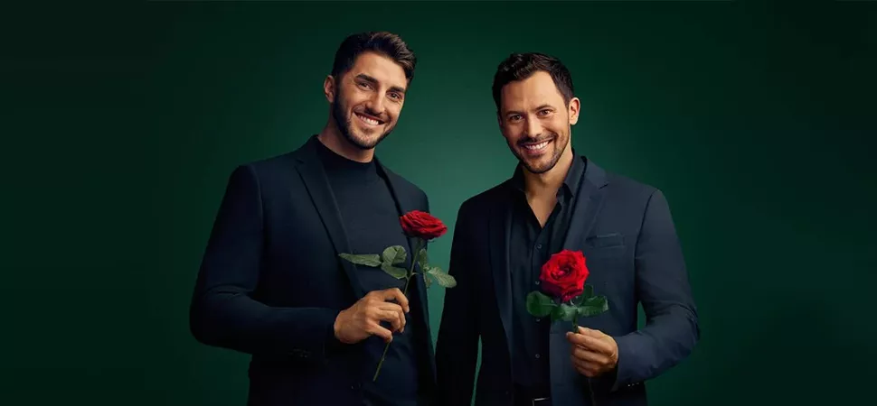 Die beiden Bachelors 2024 Dennis und Sebastian stehen im schwarzen Anzug nebeneinander und halten jeweils eine Rose in der Hand. 