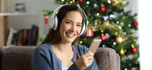 Frau mit Kopfhörern und Smartphone sitzt vor einem Weihnachtsbaum und lächelt in die Kamera