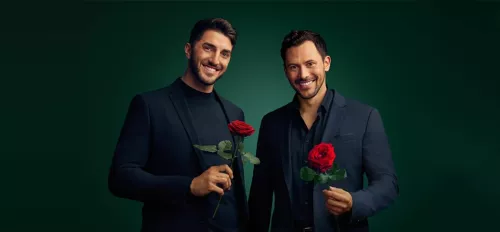 Die beiden Bachelors 2024 Dennis und Sebastian stehen im schwarzen Anzug nebeneinander und halten jeweils eine Rose in der Hand. 