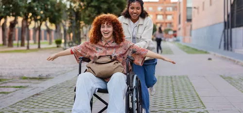 Zwei Freundinnen lachen, eine davon sitzt im Rollstuhl und streckt die Arme aus, die andere schiebt sie