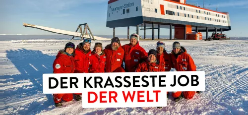 Auf dem Bild sind 8 Menschen in roten Schneeanzügen zu sehen. Sie bilden das überwinterungsteam auf der deutschen Froschungsstation Neumeyer 3 in der Antarktis. Unter ihnen ist Eisund im Hintergrund sieht man die Station.