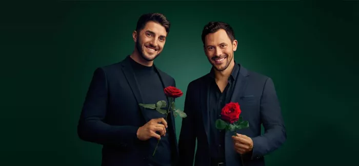 Die beiden Bachelors 2024 Dennis und Sebastian stehen im schwarzen Anzug nebeneinander und halten jeweils eine Rose in der Hand. 