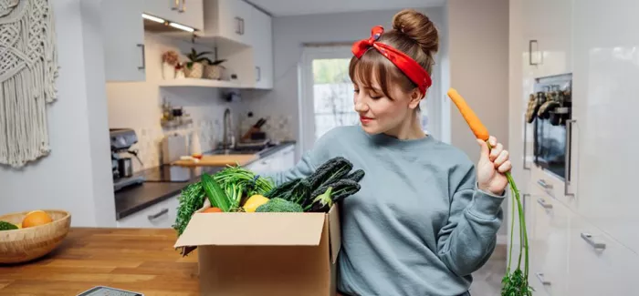 Frau mit einer Kiste voller Gemüse & einer Karotte in der Hand