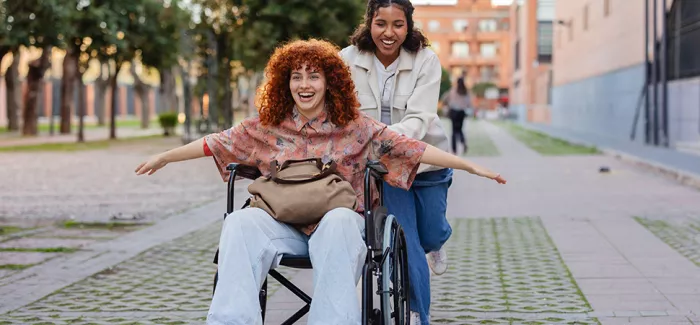 Zwei Freundinnen lachen, eine davon sitzt im Rollstuhl und streckt die Arme aus, die andere schiebt sie