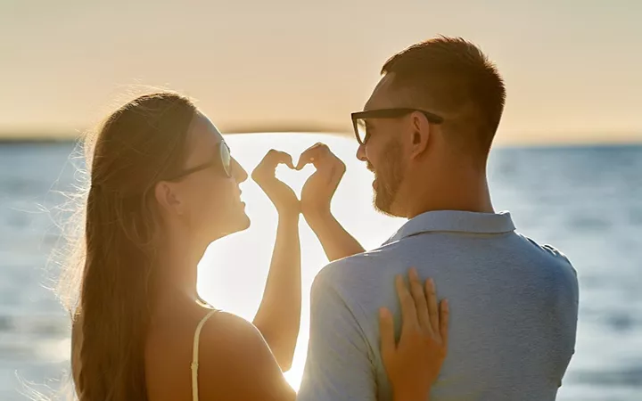 Ein Paar am Strand blicken in den Sonnenuntergang und formen ihre Hände zu einem Herz