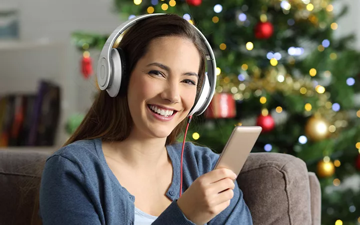 Frau mit Kopfhörern und Smartphone sitzt vor einem Weihnachtsbaum und lächelt in die Kamera