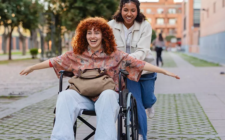 Zwei Freundinnen lachen, eine davon sitzt im Rollstuhl und streckt die Arme aus, die andere schiebt sie