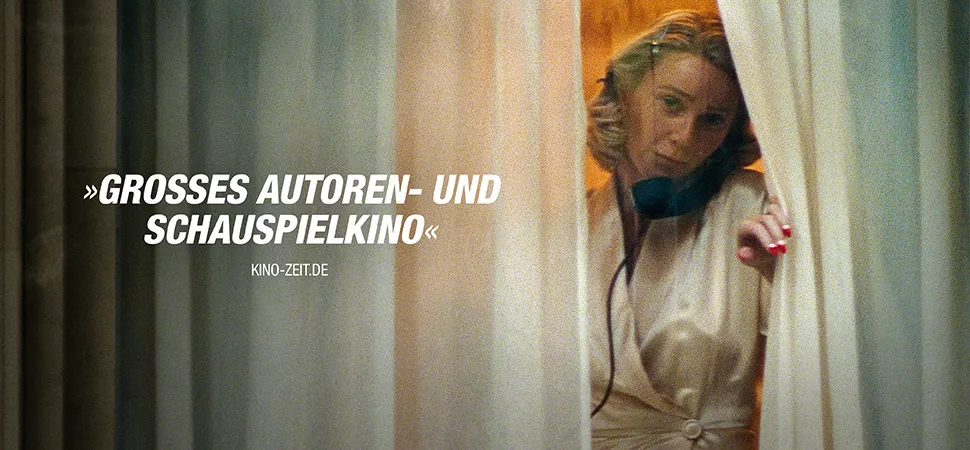 Eine telefonierende Frau schiebt einen Vorhang zur Seite und schaut aus dem Fenster